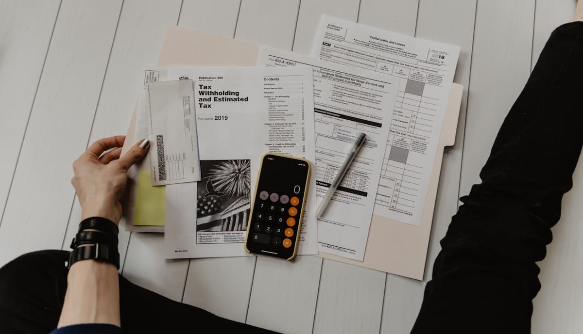 Tax forms and IRS publications spread on a wooden floor next to a phone calculator app and pen