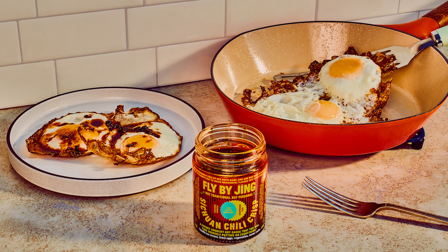 A Fly By Jing Sichuan chili crisp jar on a kitchen counter beside fried eggs on a plate and in a skillet