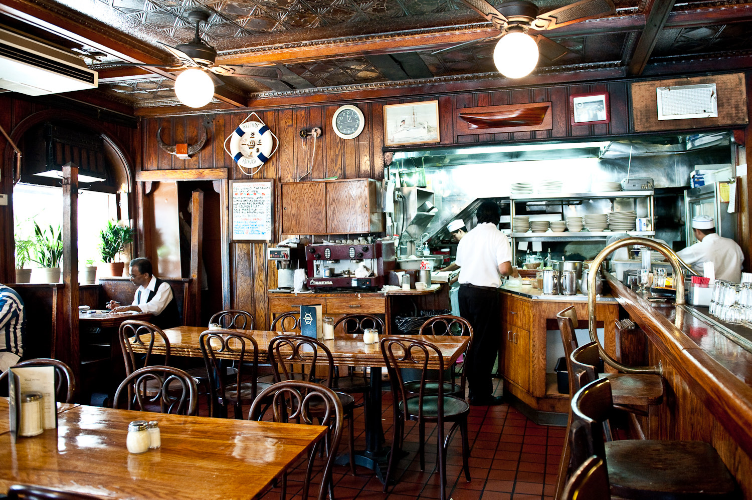 A wood-paneled restaurant dining room with an open kitchen, where cooks work the line and tables sit ready in the foreground