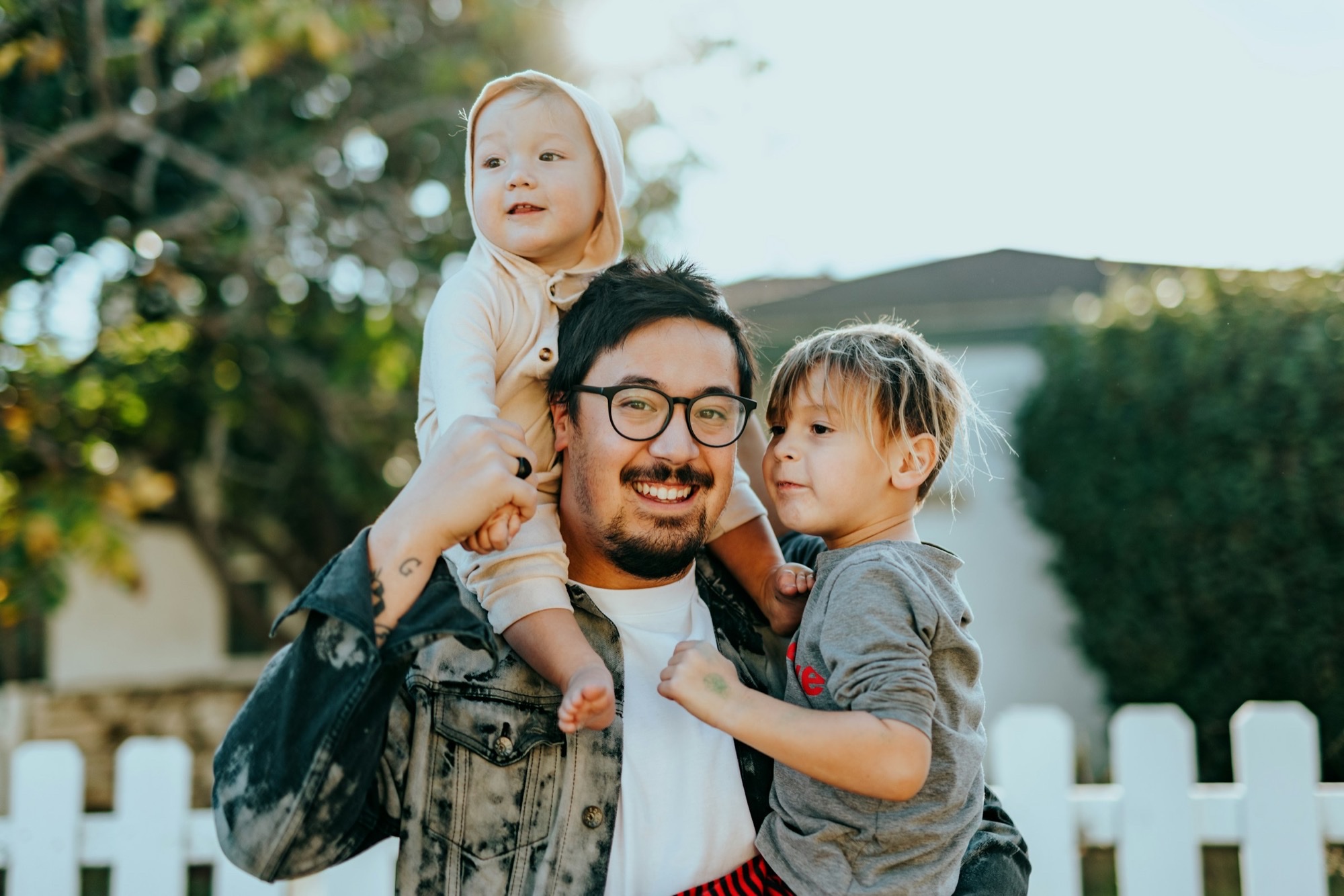 Asian American father outdoors holding two young children, smiling in soft evening light