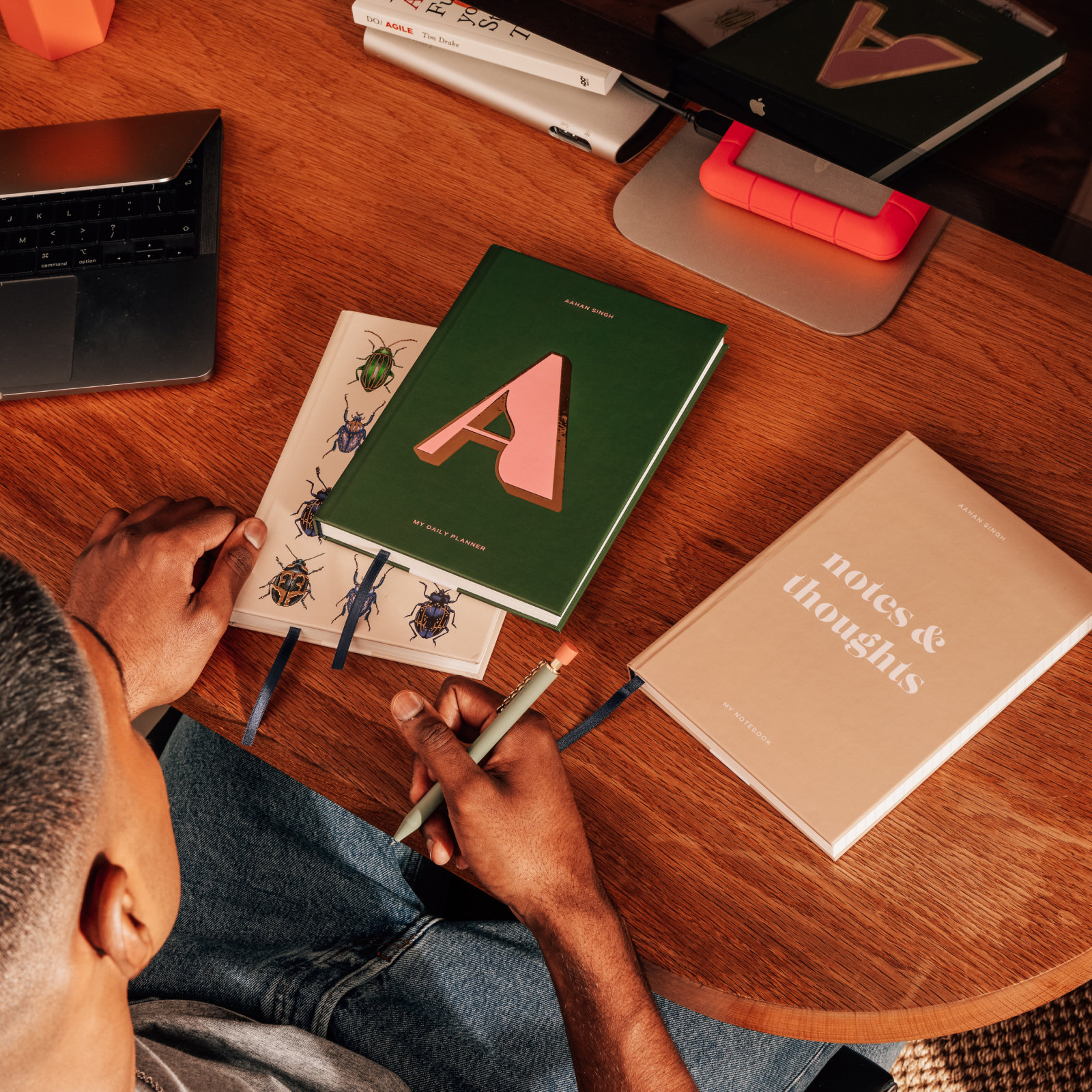 Hands arranging personalized Papier daily planners and notebooks on a wooden desk beside a laptop