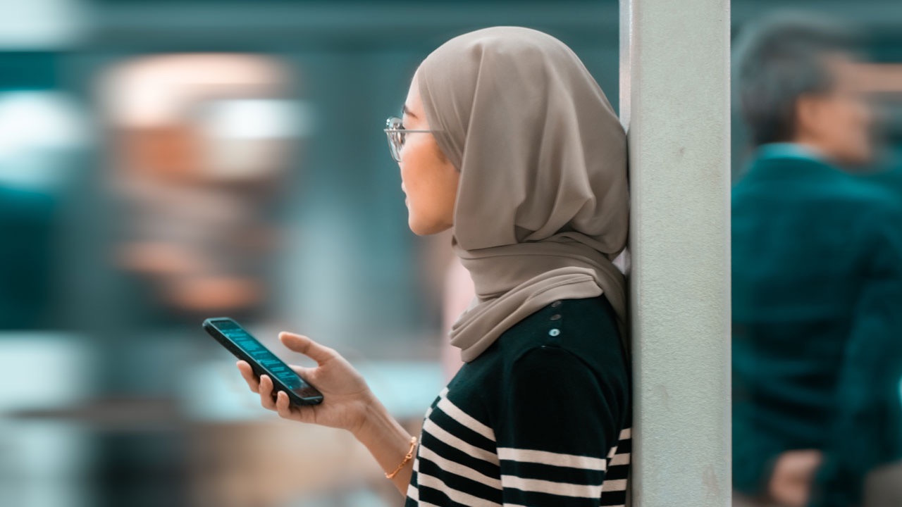 A woman in a hijab holds a smartphone, the same device used for family calls and remittance apps