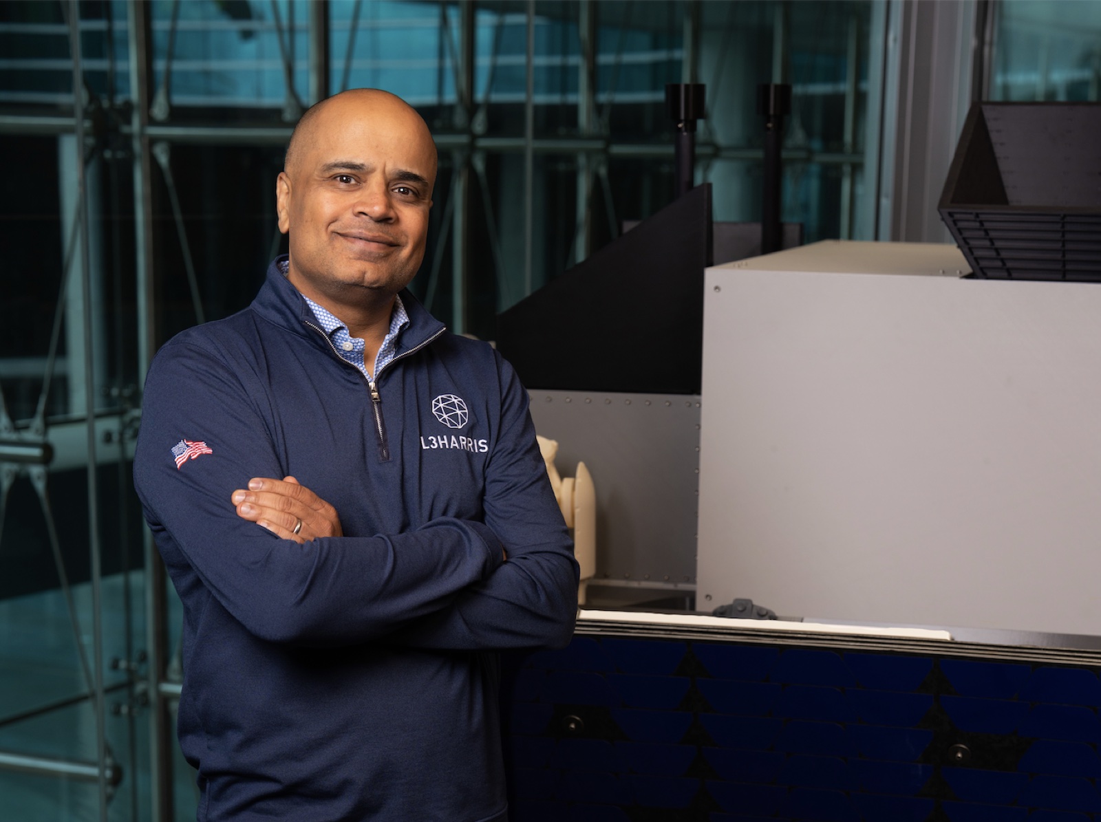 Sam Mehta, president and CEO of L3Harris Technologies, standing with arms crossed beside aerospace equipment in an L3Harris pullover